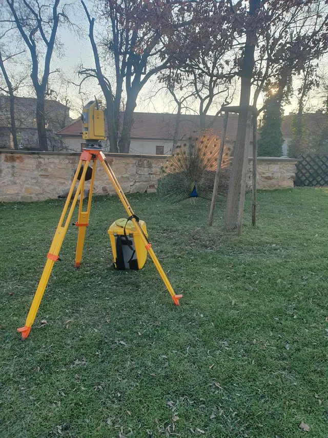 Totální stanice na stativu na zahradě, v pozadí páv s rozevřeným ocasem