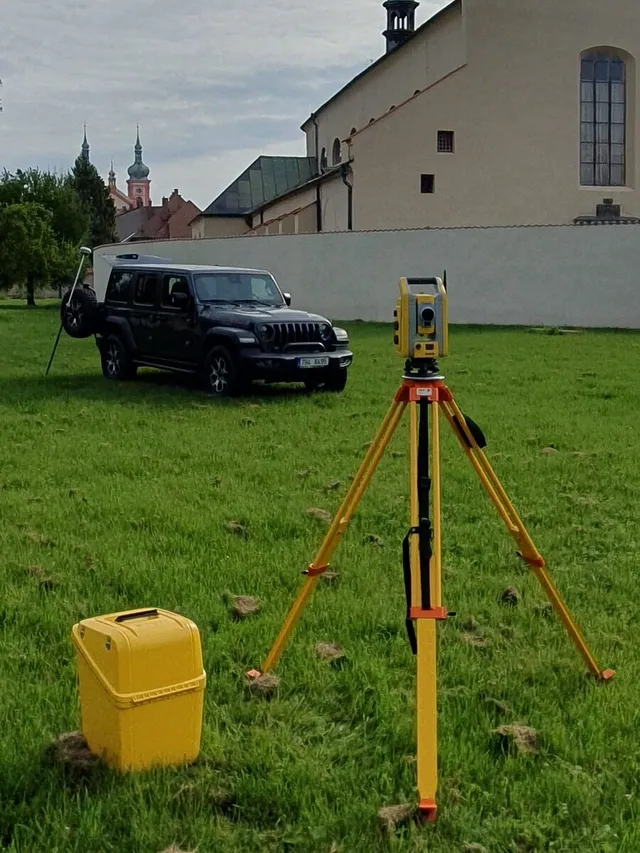 Totální stanice na stativu a terénní vůz na louce, v pozadí kostelní věž